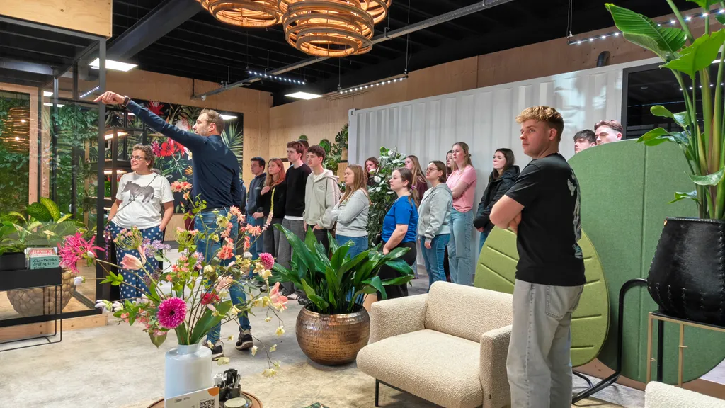 Een groep studenten krijgt uitleg in de sfeervolle showroom van Ten Brinke Interieurbeplanting, met op de voorgrond een kleurrijk zijden bloemstuk en grote kantoorplanten.