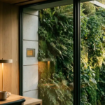 Levende groene plantenmuur op moderne gevelgevel, gezien door een kantoorraam met zacht gouden middaglicht op de bladeren.